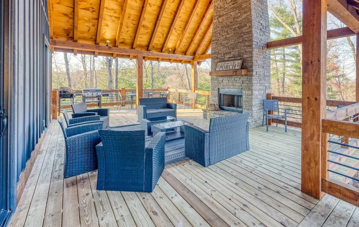 A covered wooden deck features a stone fireplace, outdoor furniture, and a railing overlooking trees under a vaulted roof.