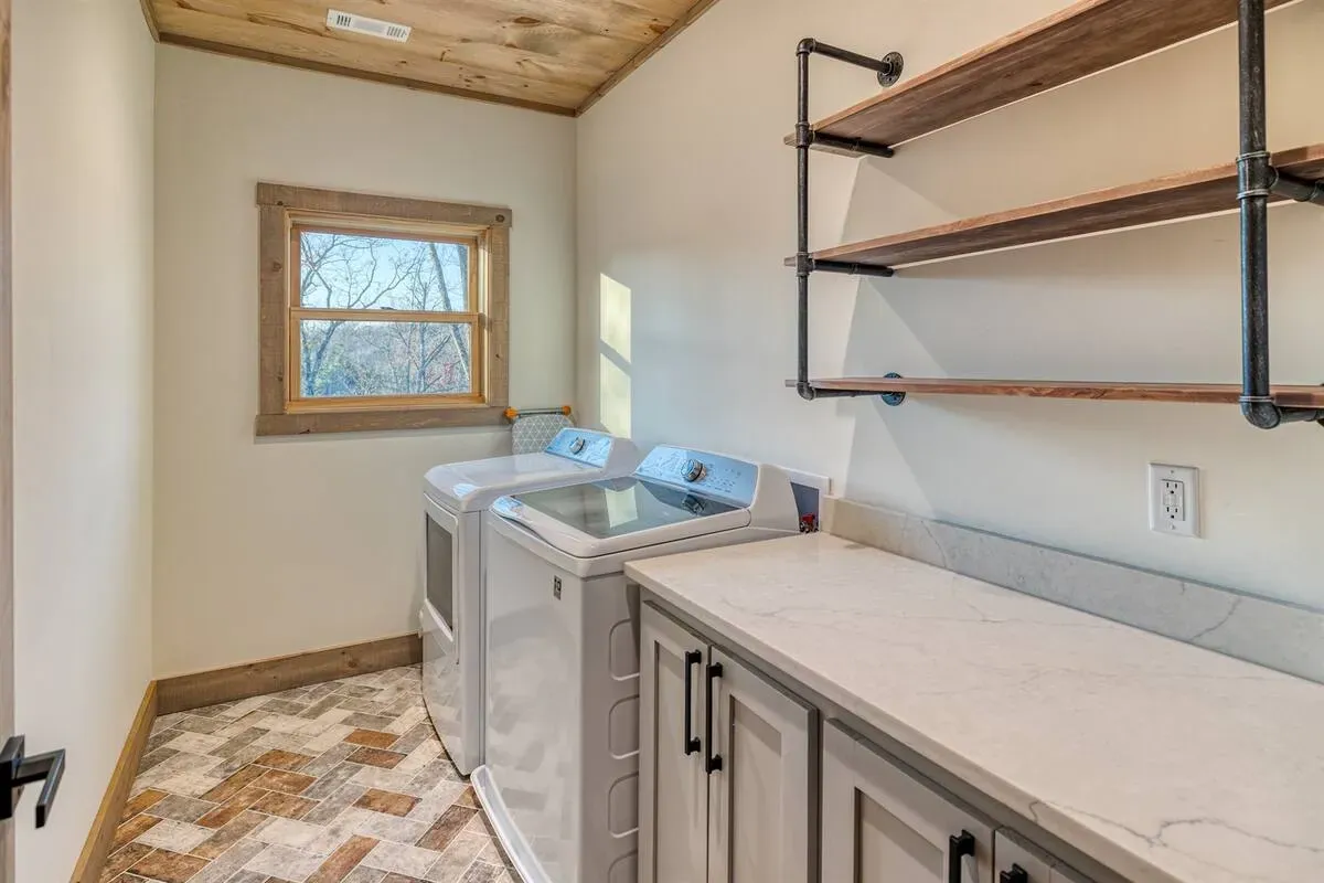 A laundry room with a washer and dryer, grey cabinets with a light countertop, and open industrial-style pipe shelving.
