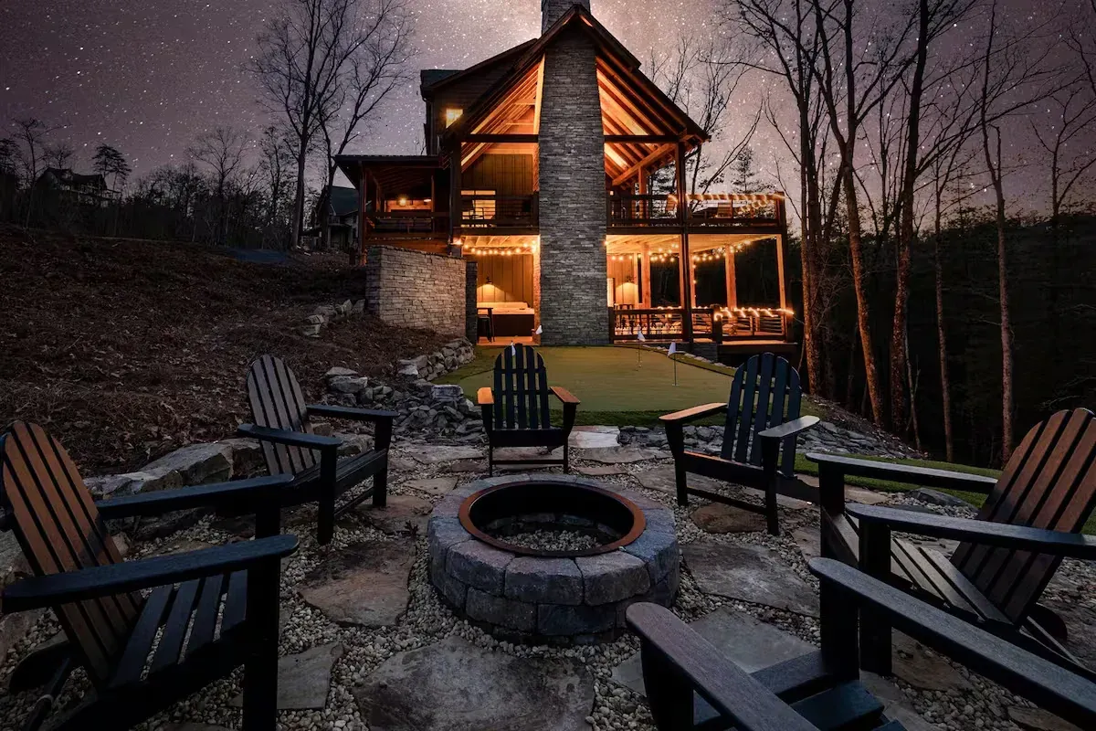 A rustic cabin at night with a stone fire pit, surrounded by black chairs and woods under a starry sky.