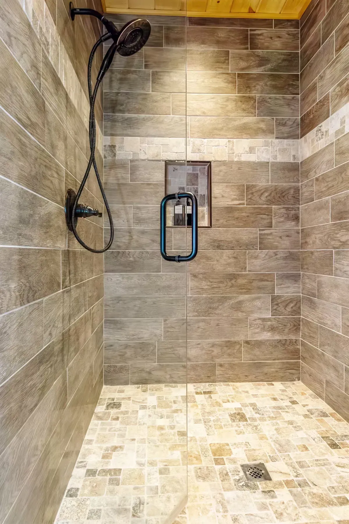 A walk-in shower with light wood-look tile walls, a mosaic stone floor, and a black handheld showerhead and door handle.