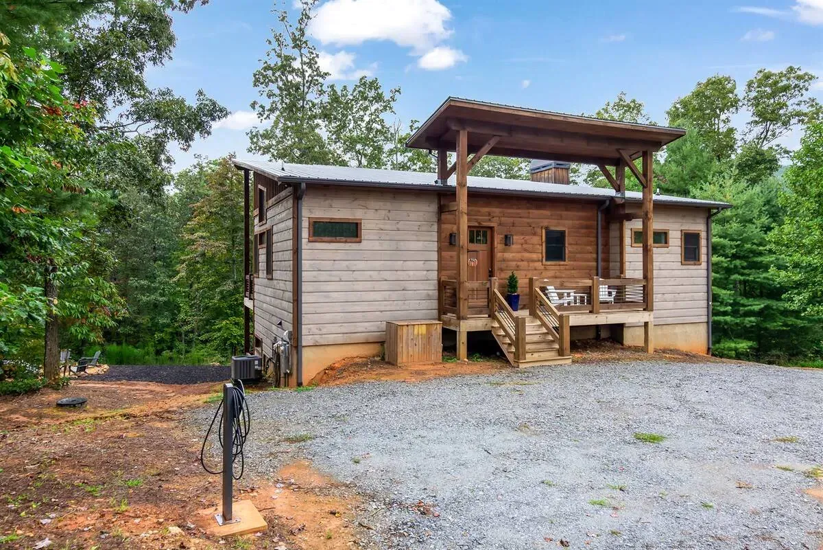 A single-story rustic cabin with wood siding, a front porch, and a pergola, set on a gravel lot surrounded by trees.