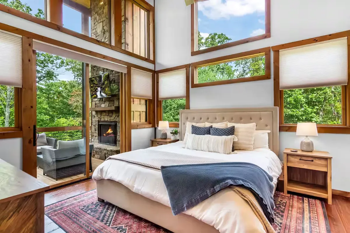 A cozy bedroom with a bed, wooden nightstand, large windows, and an outdoor stone fireplace visible through a glass door.