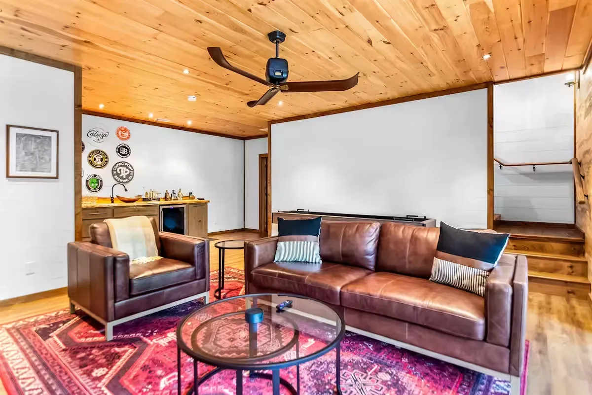 A living room with a brown leather sofa, matching armchair, circular coffee table, and patterned rug under wood ceilings.