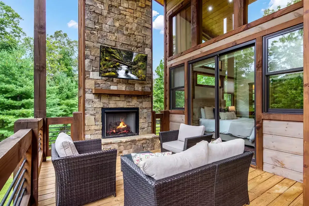 Outdoor deck with a stone fireplace, wall-mounted television, and wicker seating overlooking a wooded area.