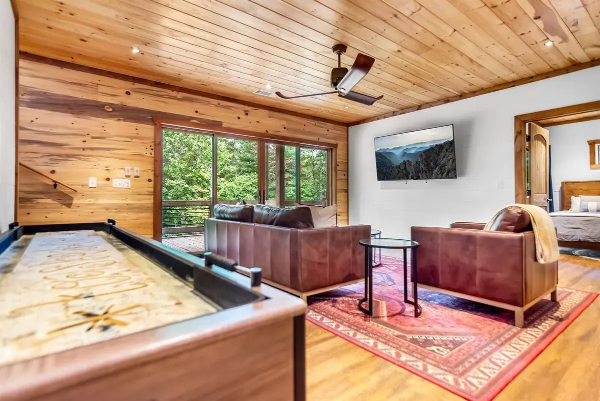 A wooden game room with a shuffleboard table, leather furniture, a patterned rug, and a wall-mounted TV.