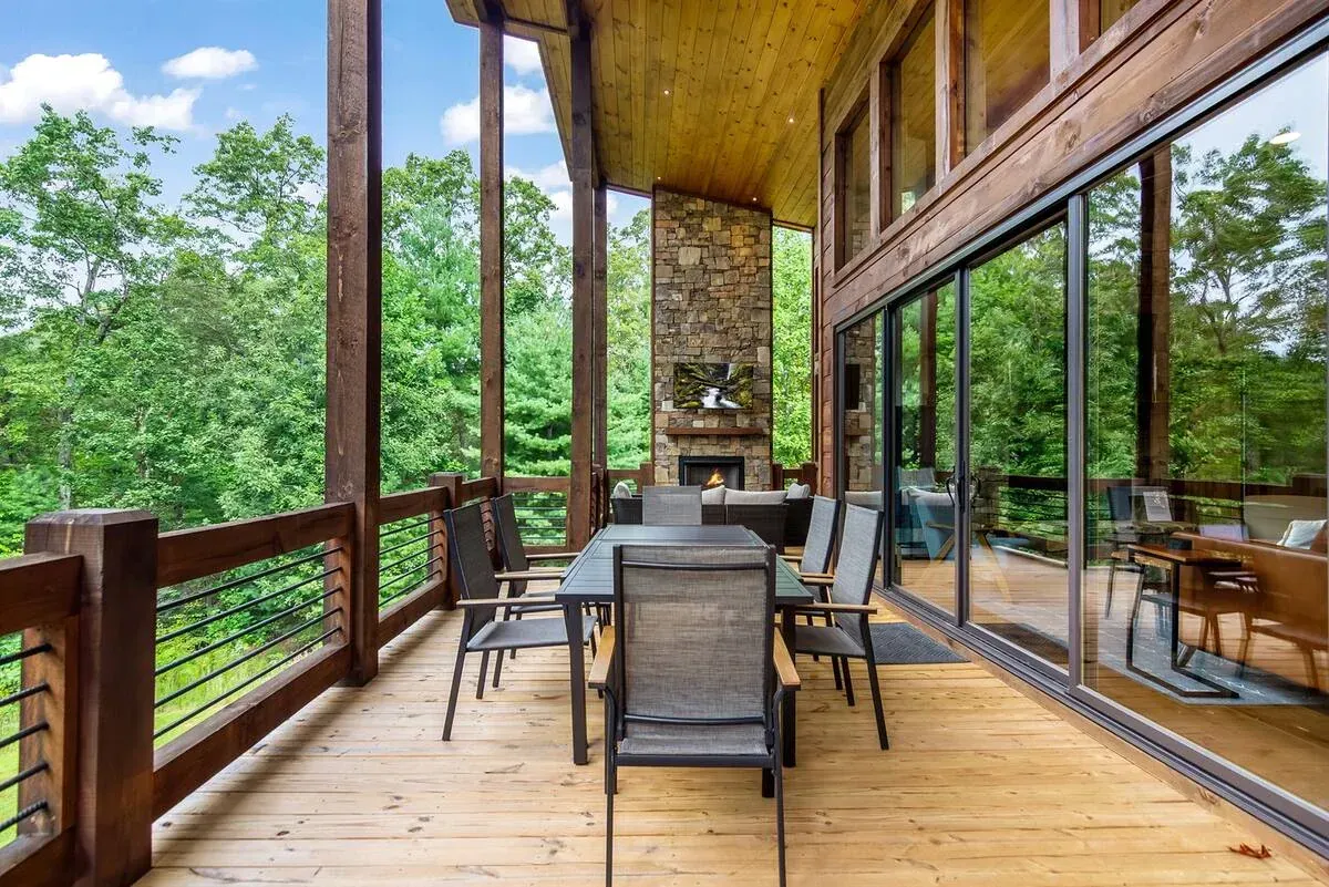 A wood-floored outdoor deck with a dining set and fireplace, overlooking a lush green forest through large glass doors.