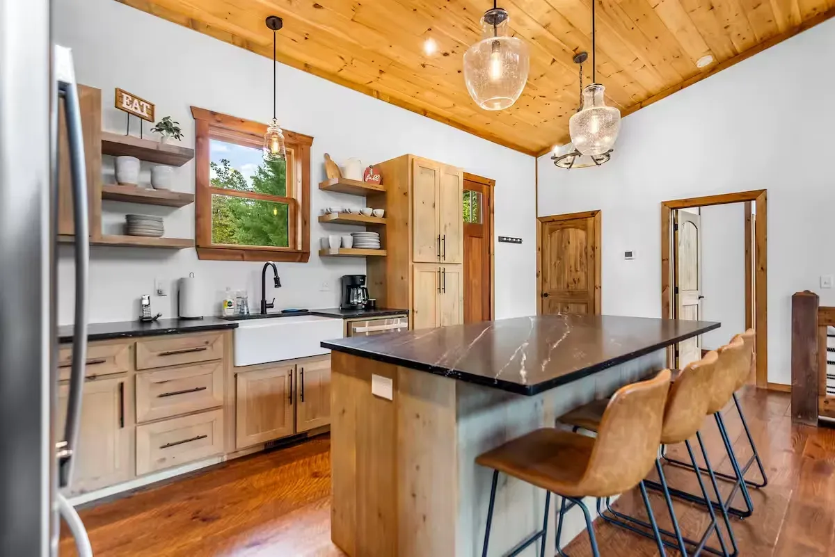 A modern, rustic kitchen features light wood cabinets, an island with dark countertops, and stools under a pine ceiling.