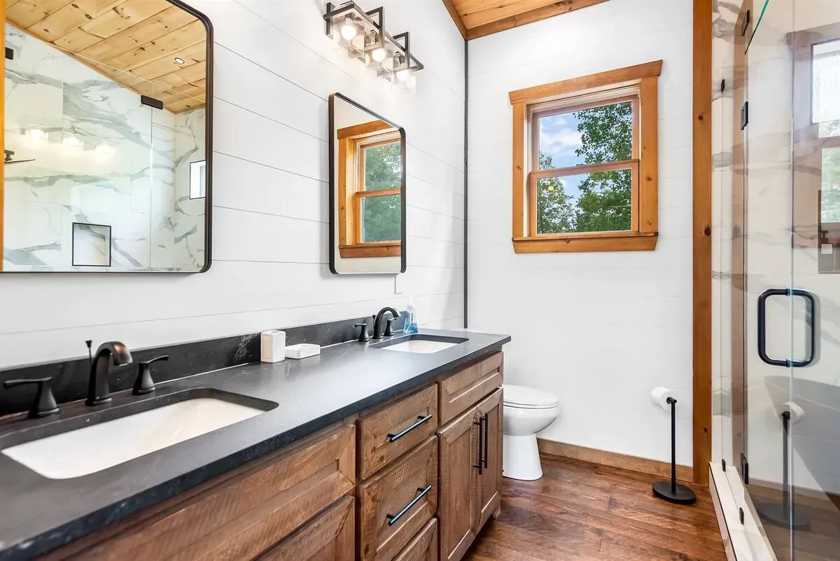 A bathroom with a double sink vanity, dark countertops, wood cabinets, white shiplap walls, and a glass-enclosed shower.