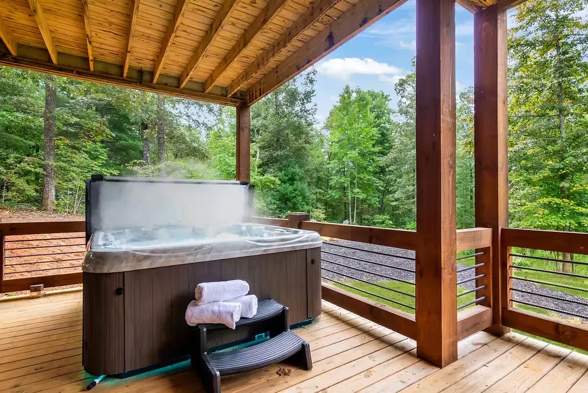 A steamy hot tub on a wooden deck overlooking a lush forest, with a stack of white towels on the steps.