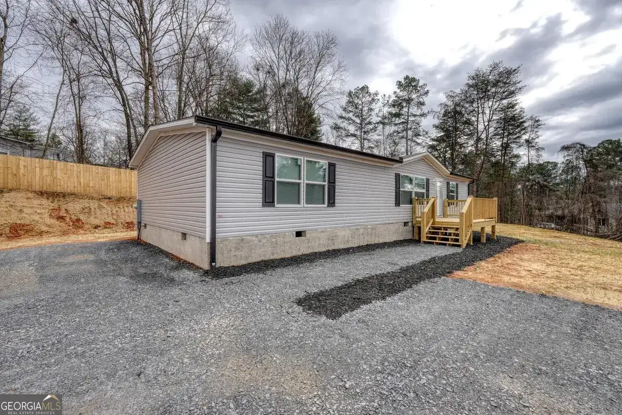 A light-colored manufactured home with a wooden deck, set on a gravel lot with a wooded background.