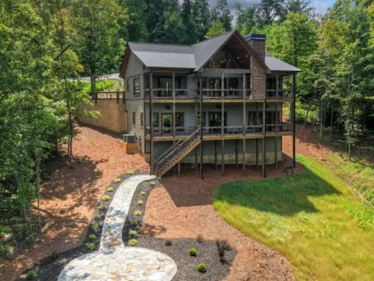 A three-story cabin with a stone chimney and wrap-around decks, surrounded by trees and a landscaped stone walkway.