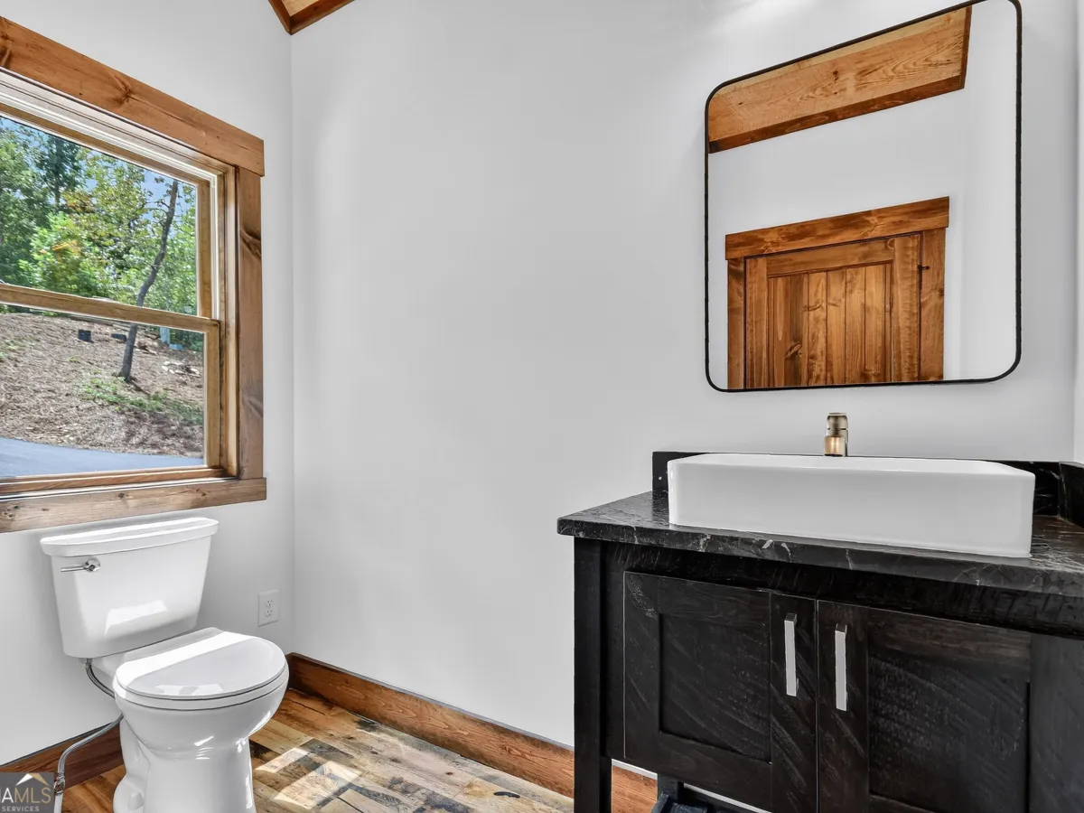 A bathroom featuring a black vanity with a white vessel sink, a rectangular mirror, and a white toilet by a wooden window.