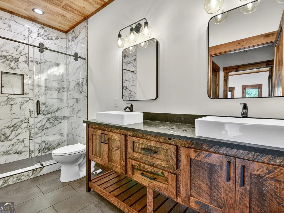 A rustic bathroom with dual white vessel sinks, dark wood vanity, marble-tiled shower, and two rectangular mirrors.