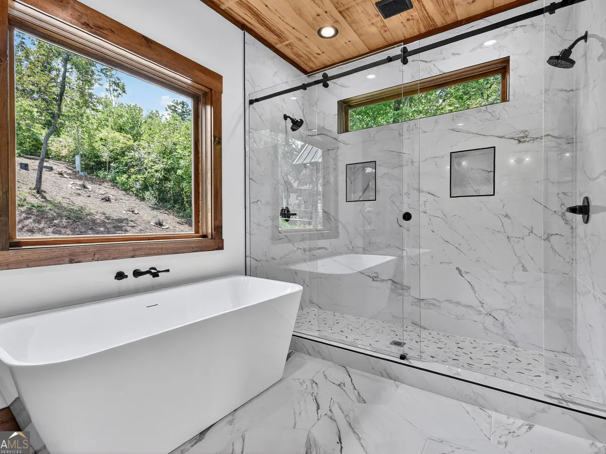 Modern bathroom with a freestanding white tub, walk-in glass shower, marble tile, and a large window facing the woods.