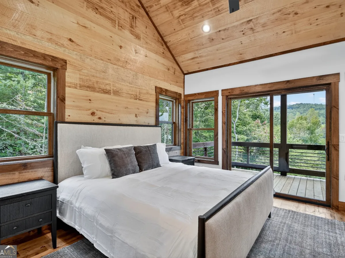 A rustic bedroom with a king bed, light wood walls, vaulted ceiling, and sliding glass doors revealing a mountain view.