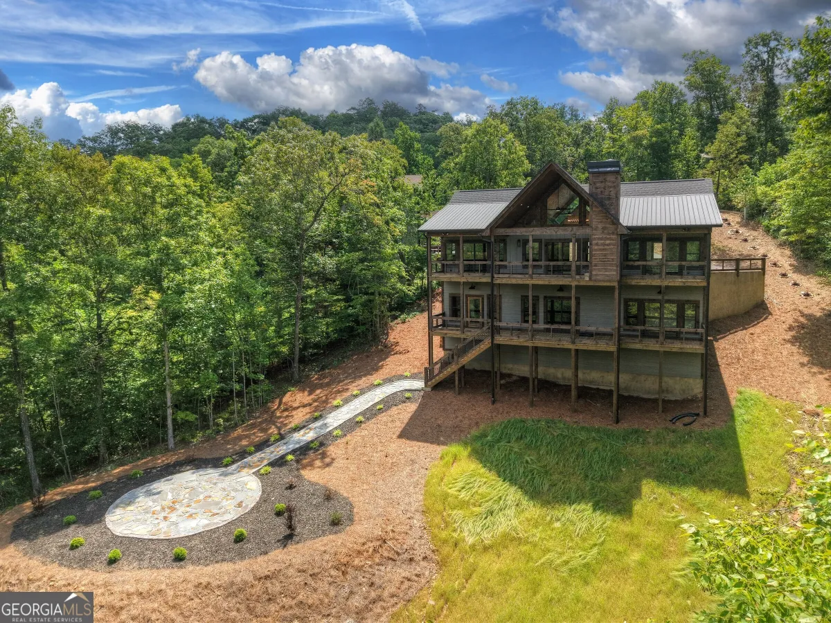 A three-story rustic wooden cabin with a metal roof nestled in a forested landscape, featuring a stone patio and walkway.
