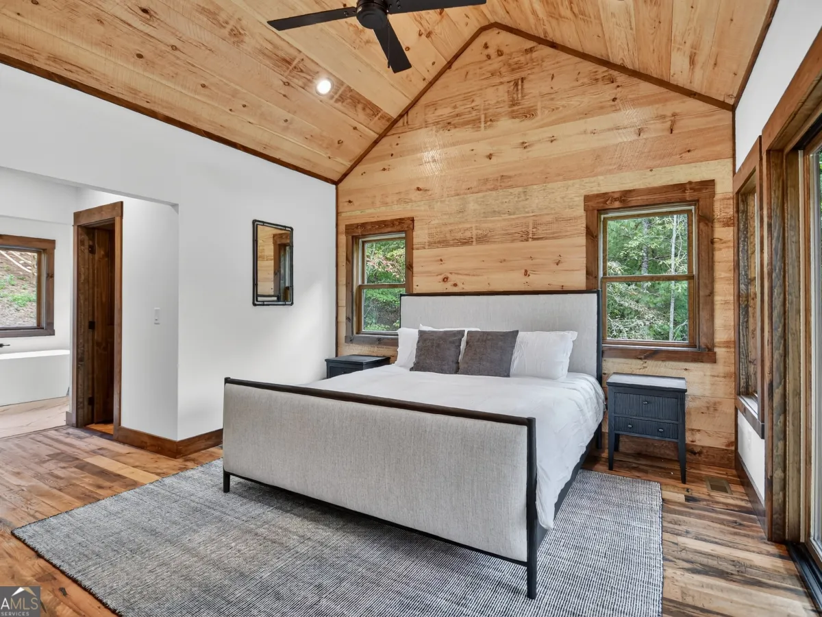 A bedroom with a queen-sized bed, white linens, two dark nightstands, a grey rug, and natural wood walls and ceiling.