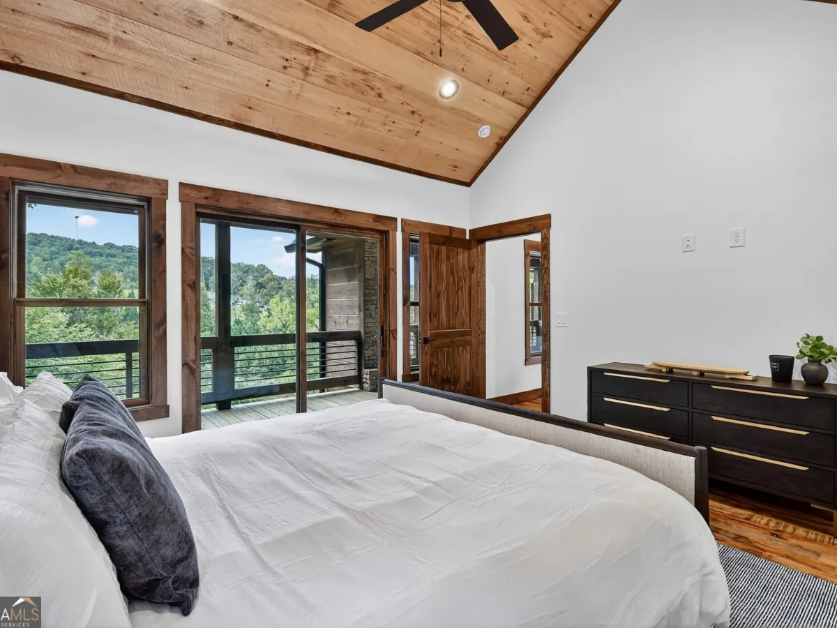 A bedroom with a vaulted wooden ceiling, white walls, a bed in the foreground, and a glass door leading to a balcony.