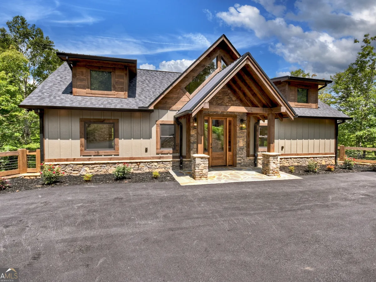 A modern mountain-style home with tan siding, stone foundation, dark roof, and a large wood-framed front porch.