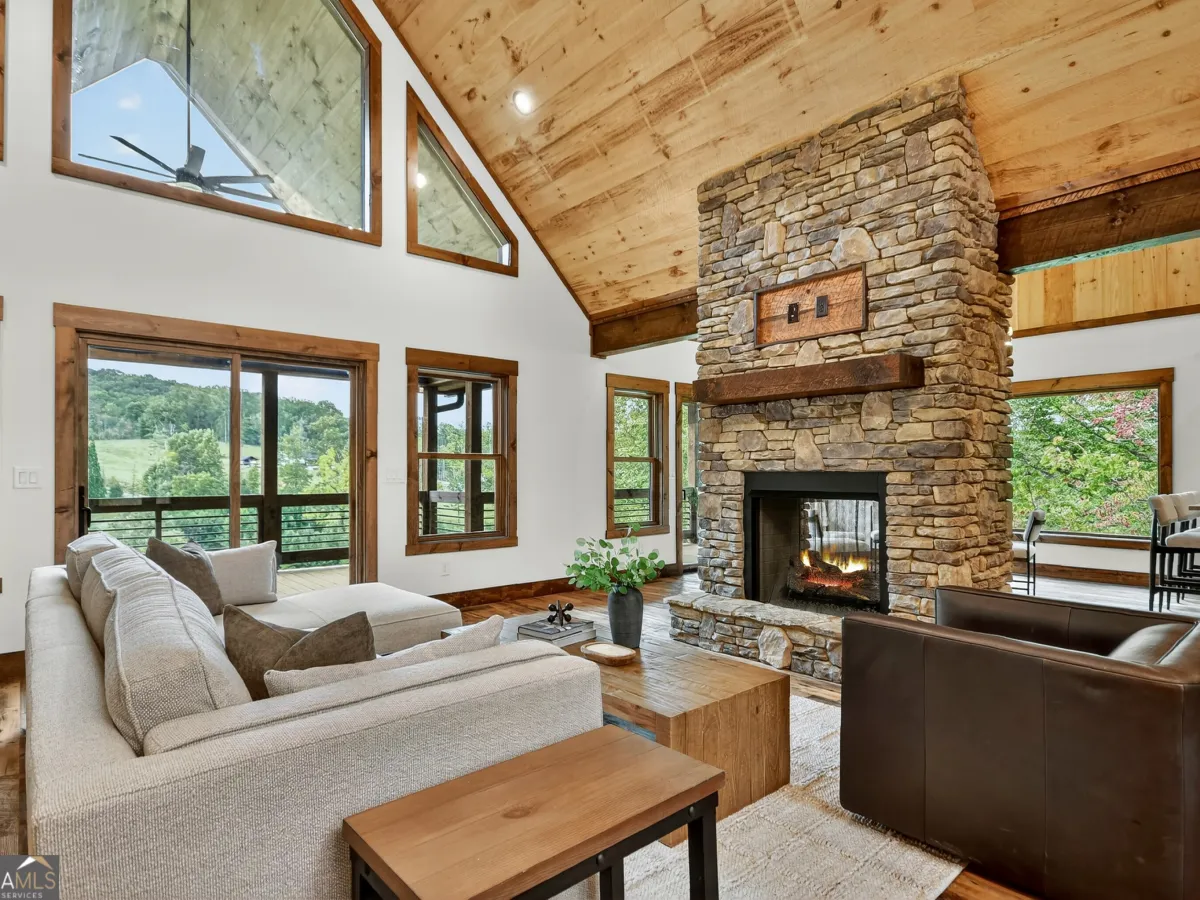 Living room with a stone fireplace, high wooden ceiling, large windows, light sofa, and dark leather armchair.