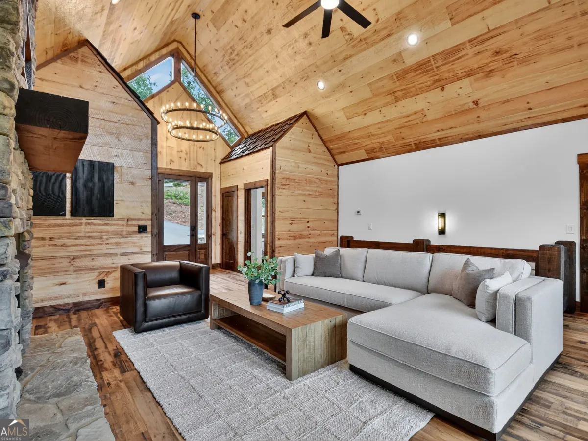 A living room with a stone fireplace, gray sectional sofa, leather armchair, wood-paneled walls, and vaulted ceilings.