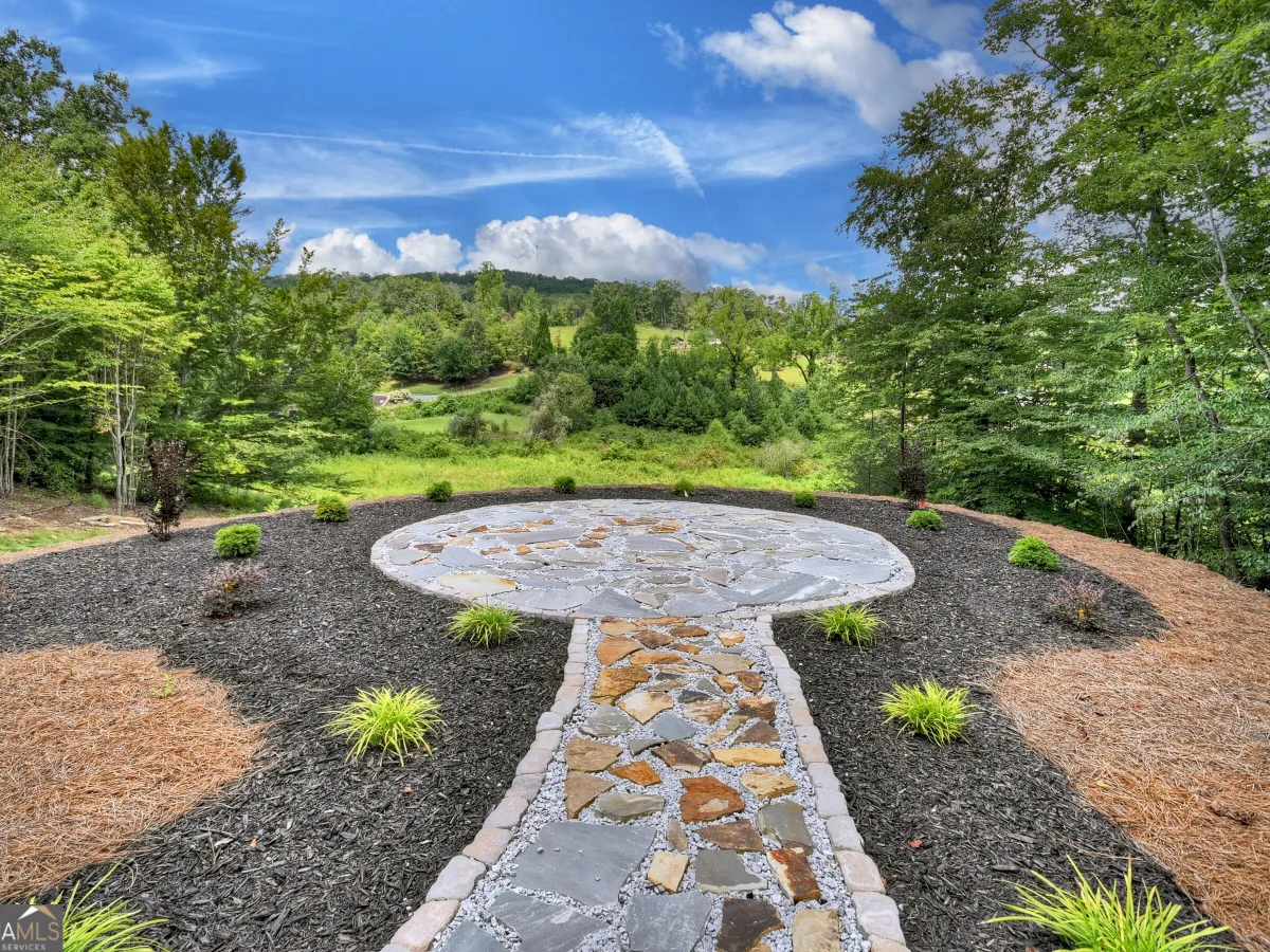 A stone path leads to a circular patio in a landscaped yard, set against a backdrop of trees and a blue sky.
