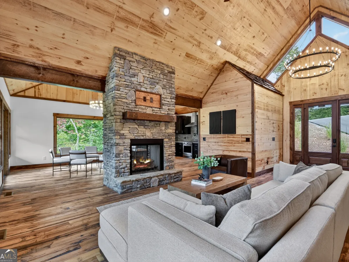 A rustic living room with wood-paneled walls, a vaulted ceiling, a large stone fireplace, and a gray sectional sofa.