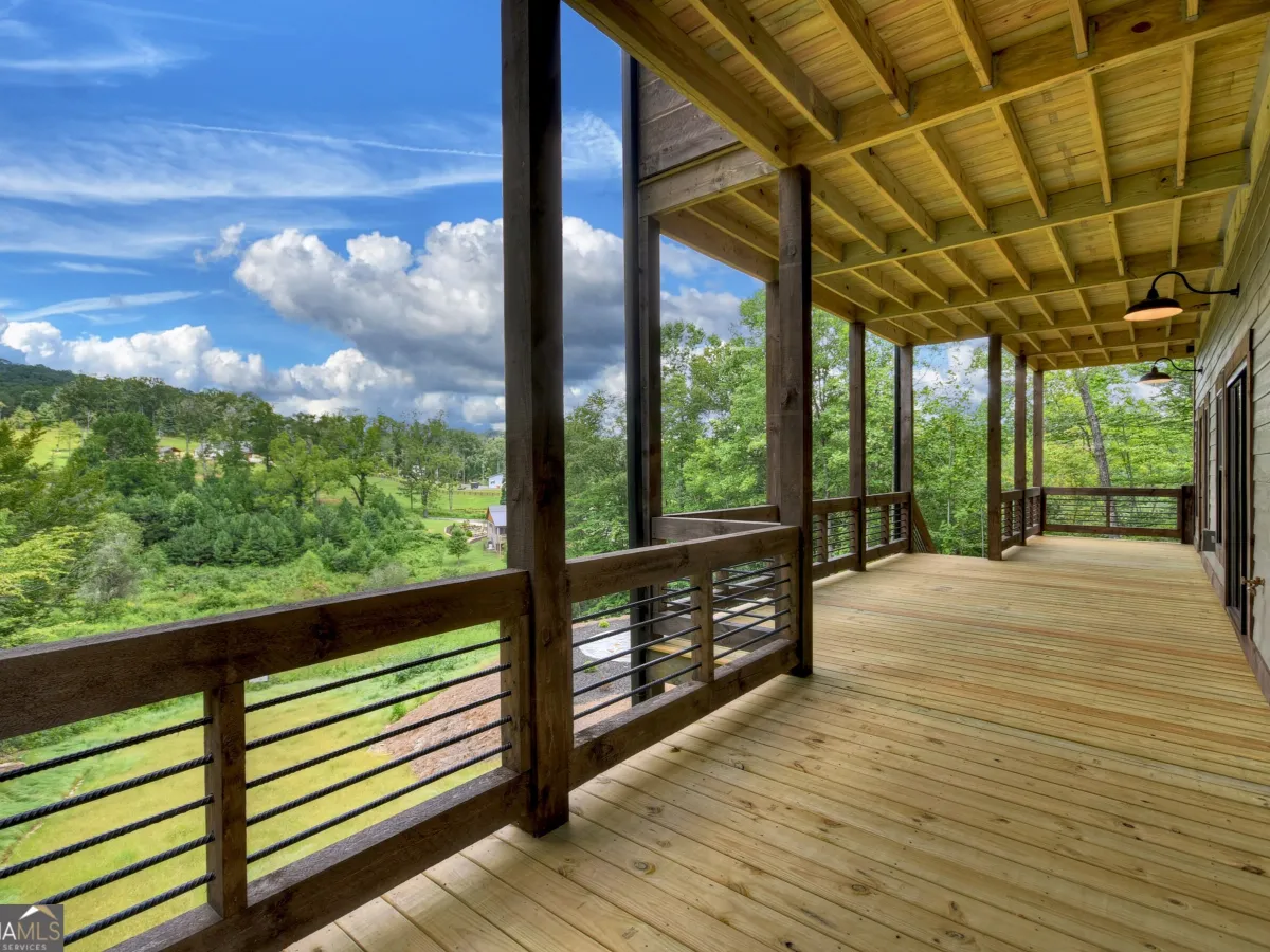 A spacious, wooden covered deck featuring cable railing and scenic views of a grassy hillside under a blue, cloudy sky.