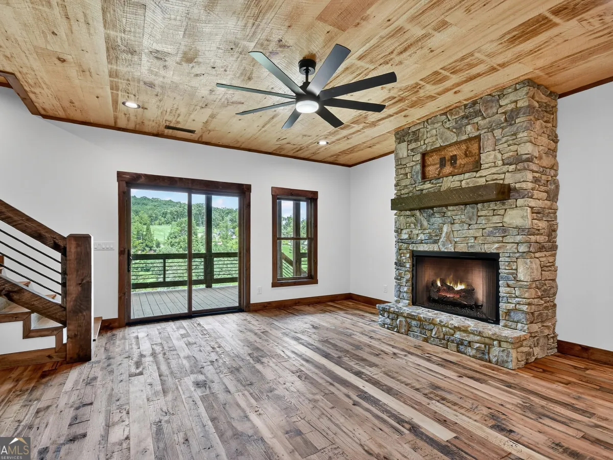 A rustic living room with wood plank walls and ceilings, stone fireplace, large sliding glass doors, and a ceiling fan.