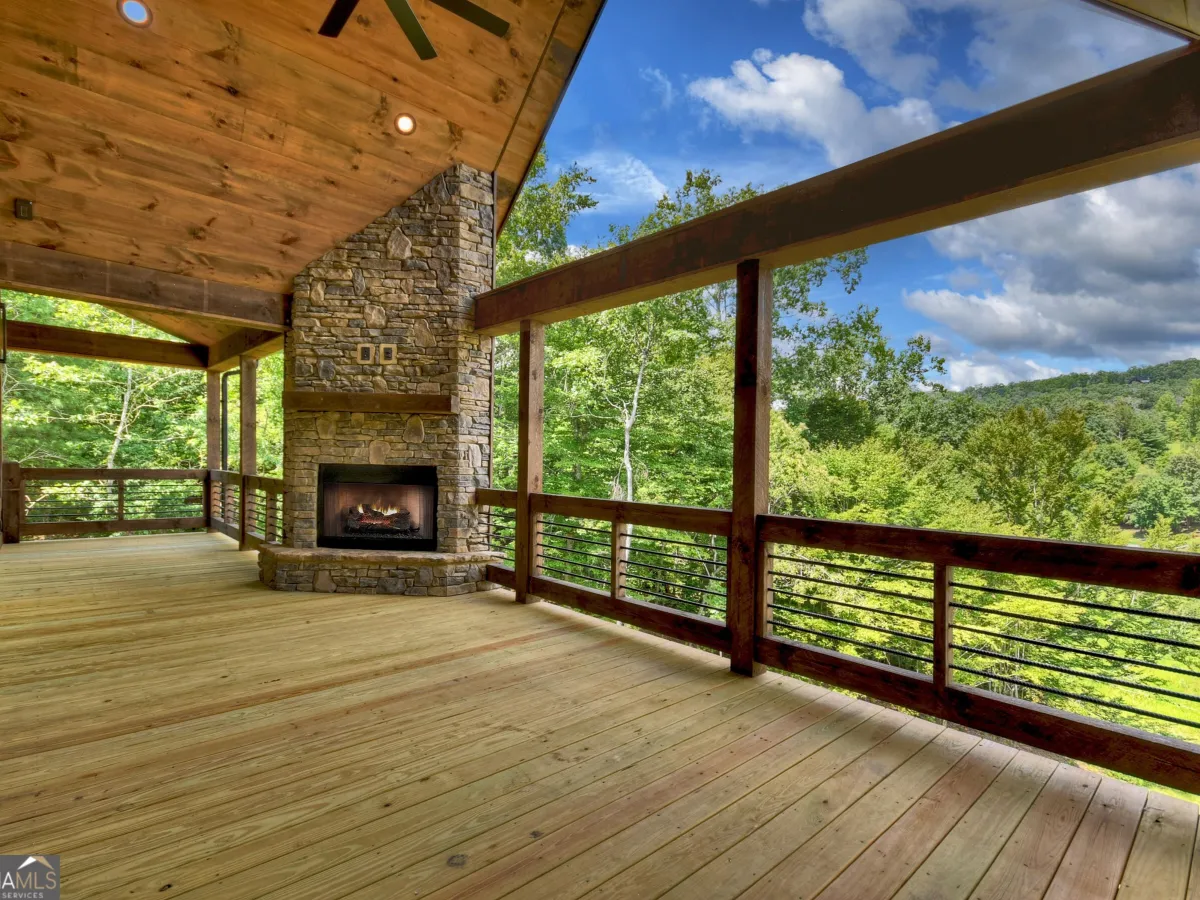 A spacious wooden deck featuring a stone fireplace, under a peaked roof, overlooking a lush, mountainous forest landscape.