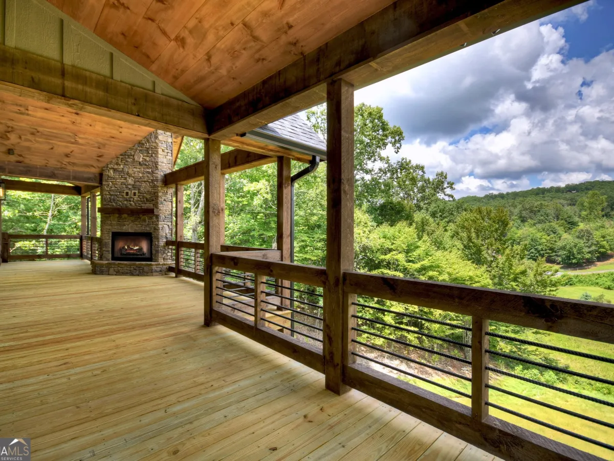 A wooden deck with a stone fireplace, overlooking a lush green valley under a bright, cloudy sky.