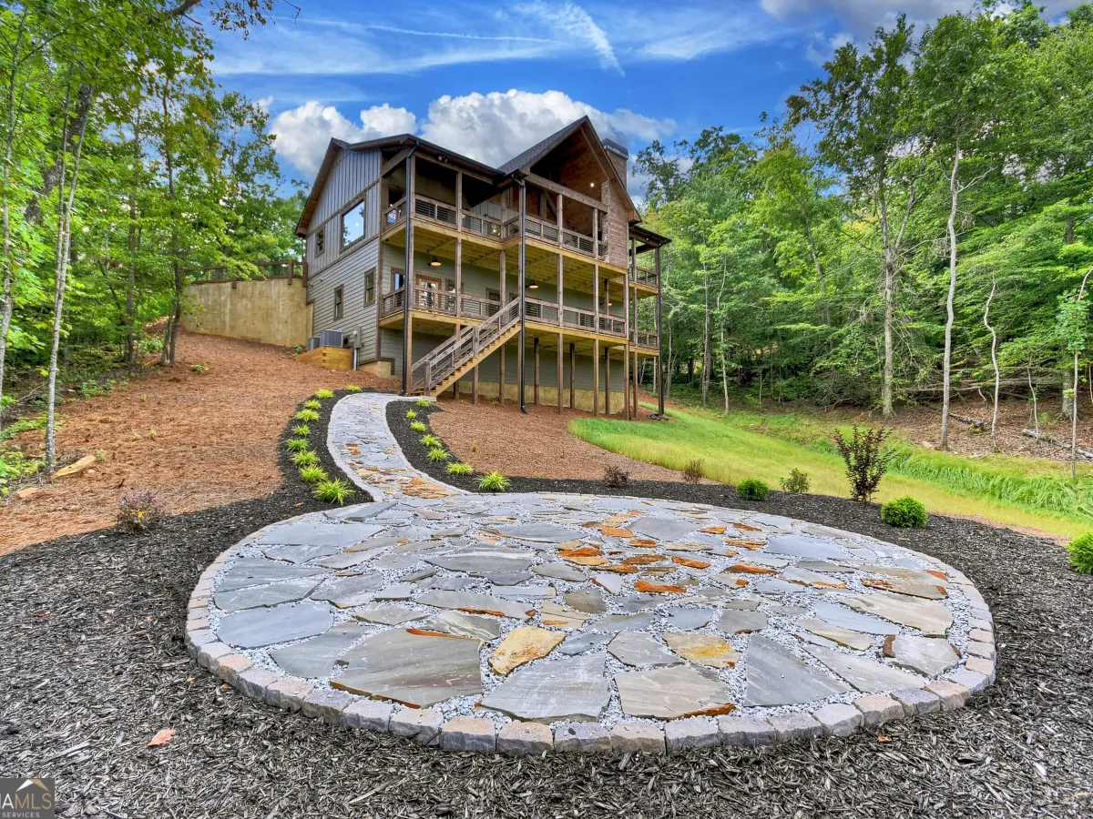 A three-story cabin nestled in a wooded landscape with a circular flagstone patio and winding path in the foreground.