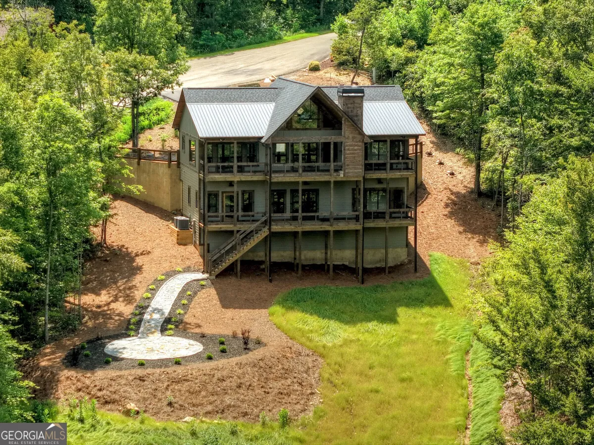 Aerial view of a three-story, grey cabin with a metal roof nestled in a forest with a circular stone patio.