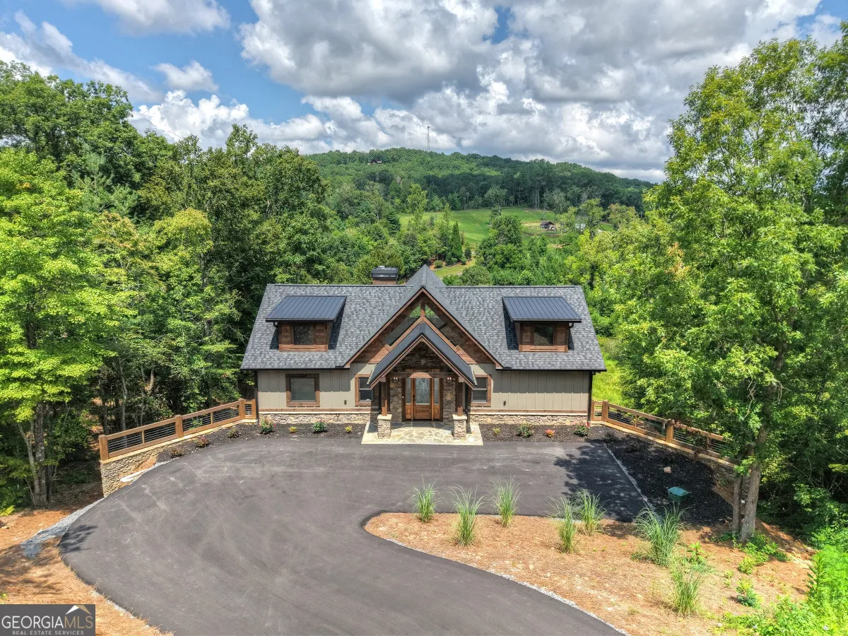 A light-colored house with a dark roof surrounded by lush green trees, set behind a curved asphalt driveway.