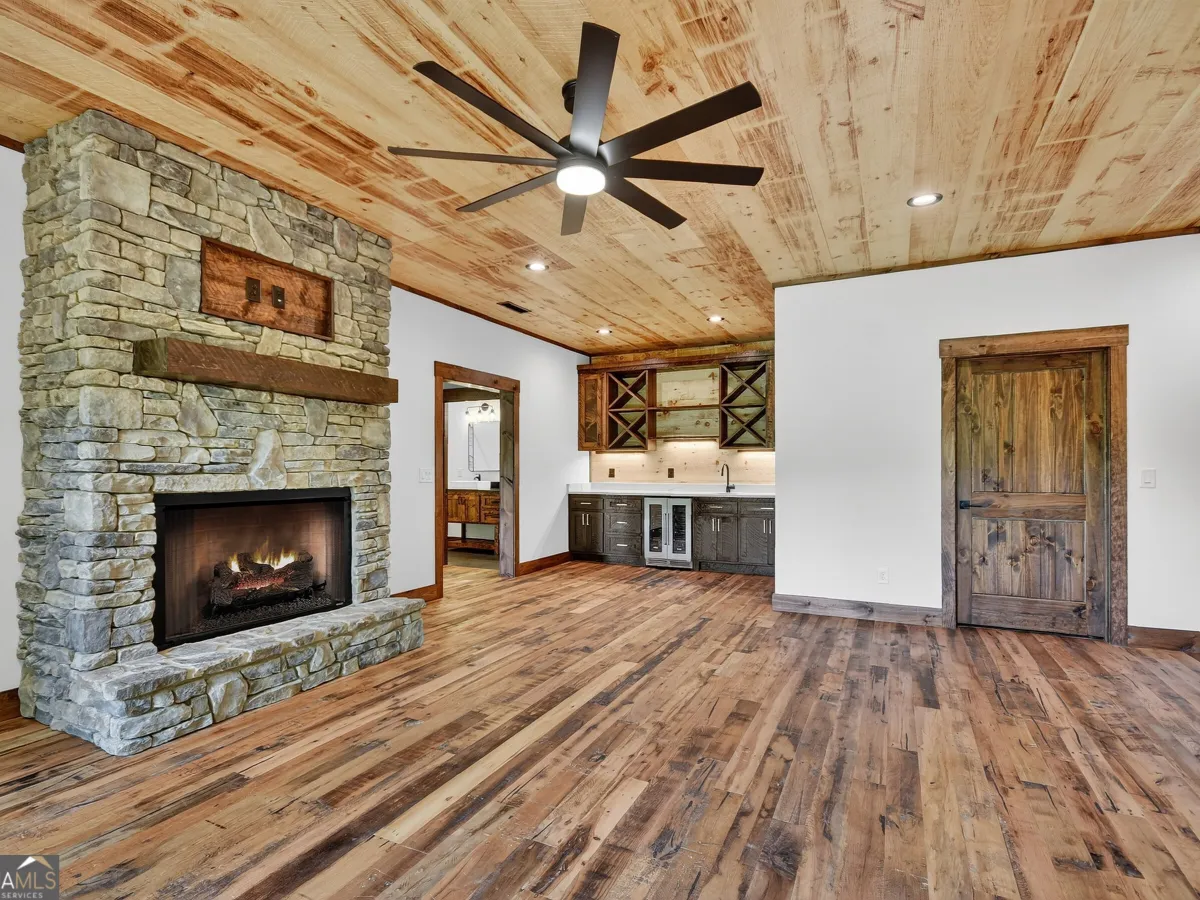 A rustic living room with wood-paneled ceiling and floors, a stone fireplace, and a kitchenette in the background.