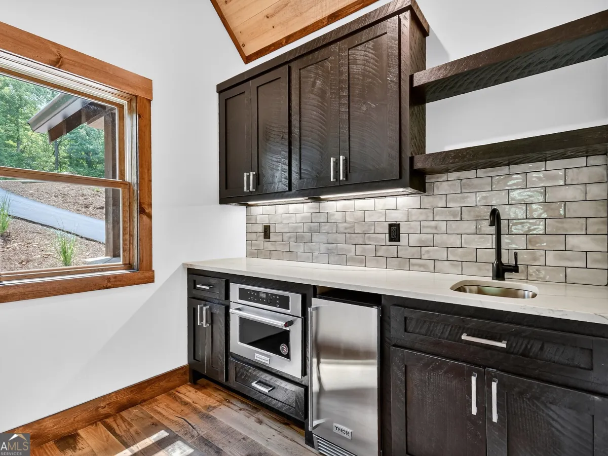 Modern wet bar with dark wood cabinets, a stainless steel microwave and mini fridge, subway tile, and a window view.
