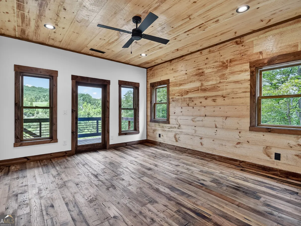 A room with wood-paneled walls and ceiling, hardwood floors, a ceiling fan, and large windows looking onto green trees.
