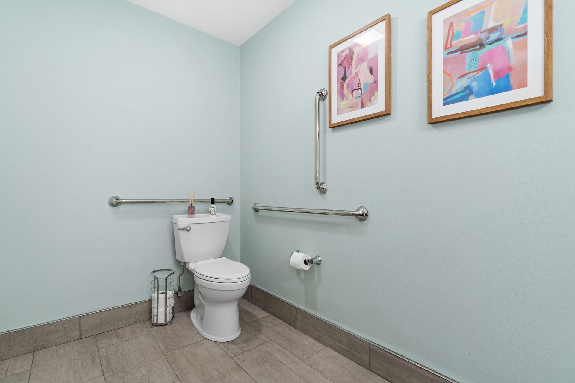 A bathroom with a toilet , handrails , and two paintings on the wall.