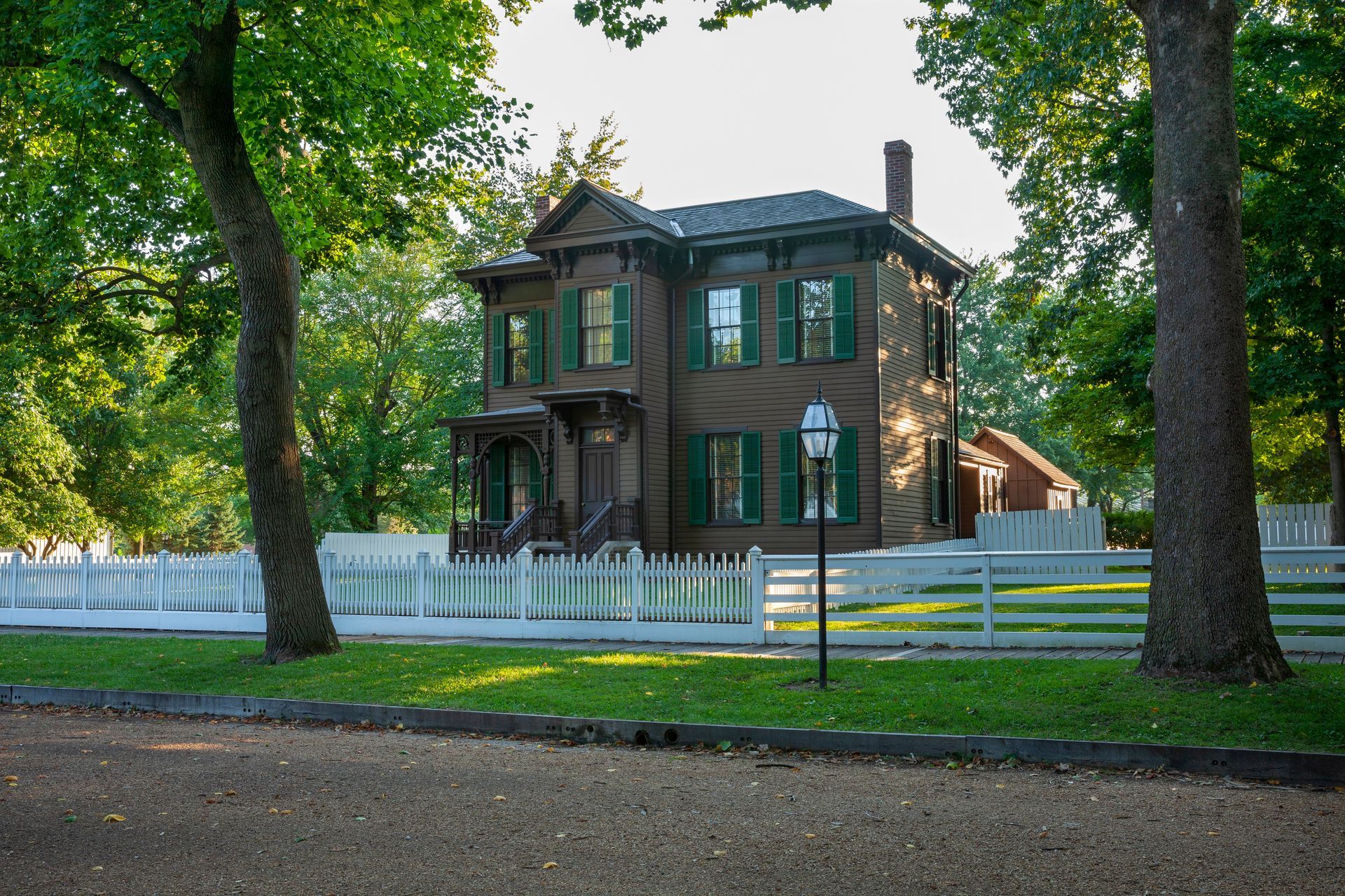 Brown house with green shutters and white picket fence, surrounded by trees and grass.