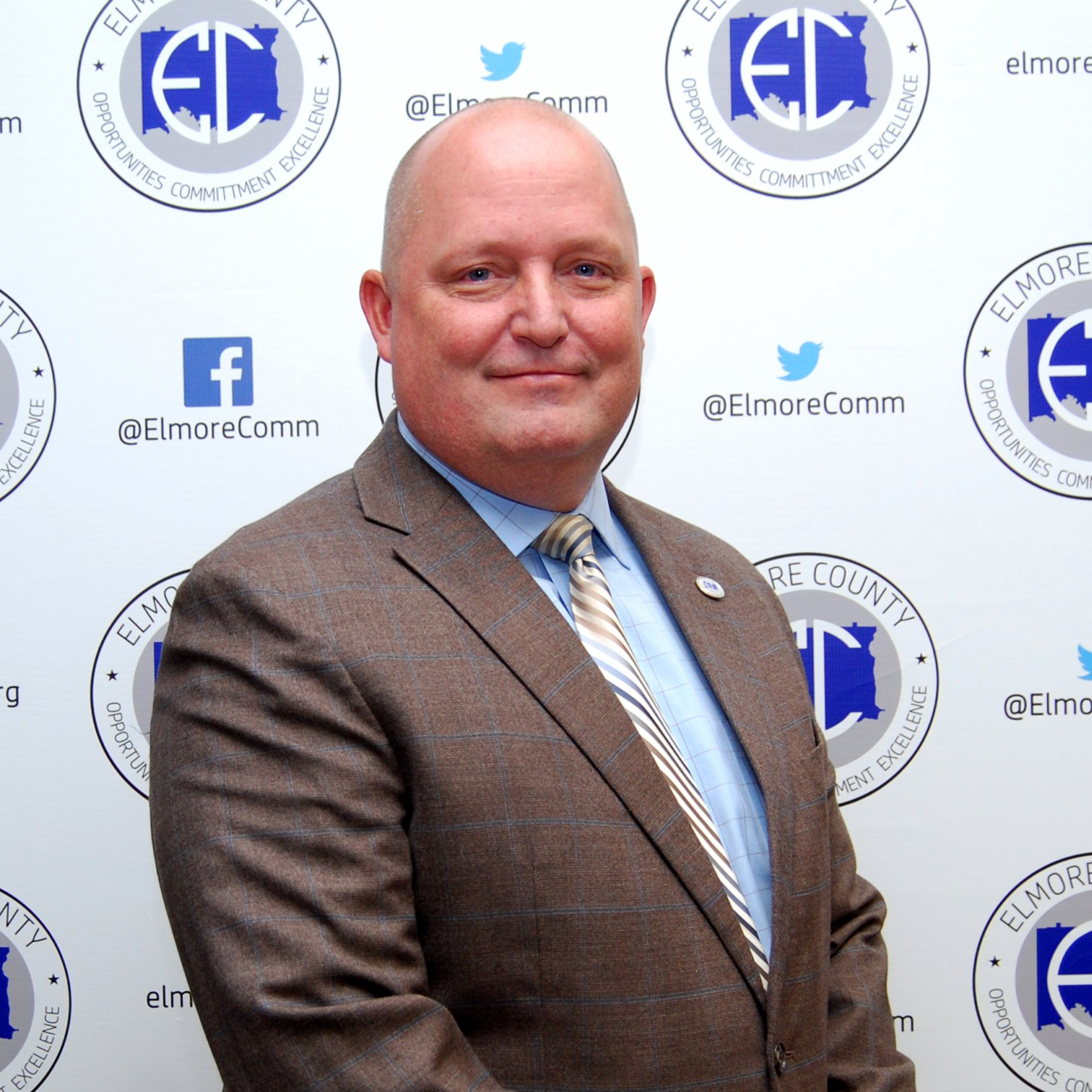 A middle-aged, bald man in a suit and tie stands in front of a backdrop with the