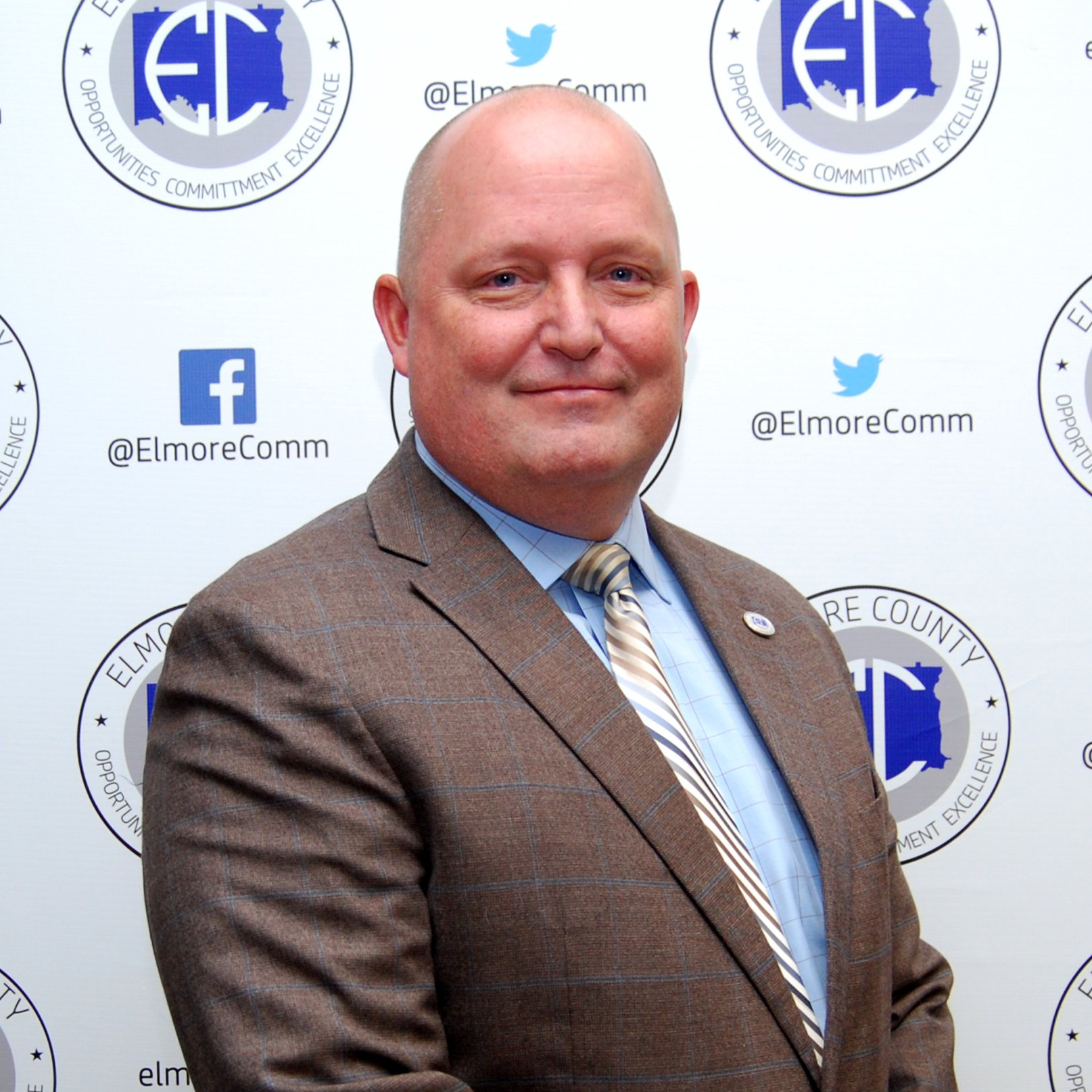 Man in suit, light blue shirt, patterned tie; Elmore County logo background.