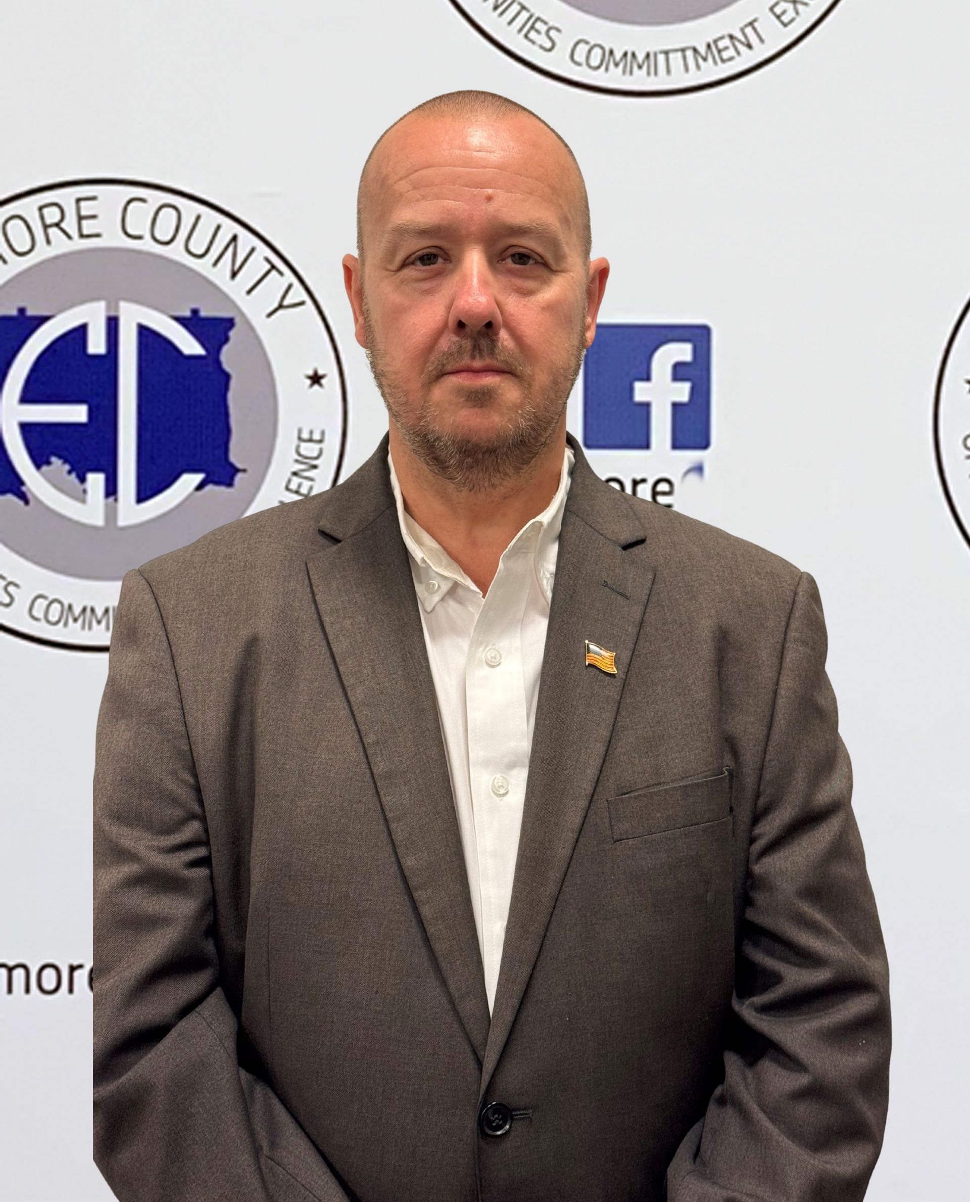 Man in a brown blazer and white shirt, standing in front of a backdrop with logos.