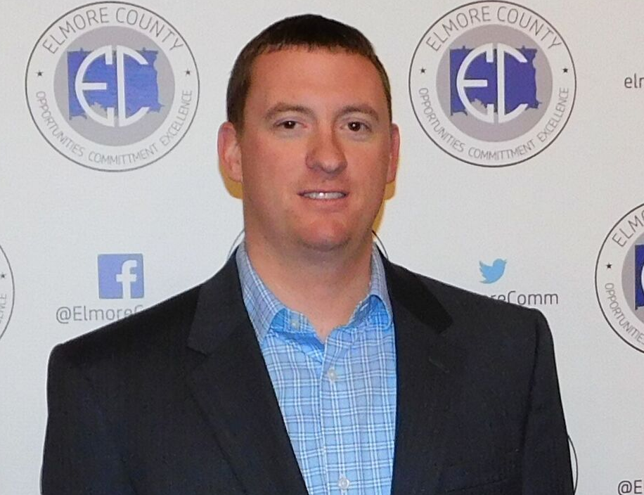 A man with short brown hair smiles, wearing a blue plaid shirt and navy blazer, in front of an Elmore County backdrop.