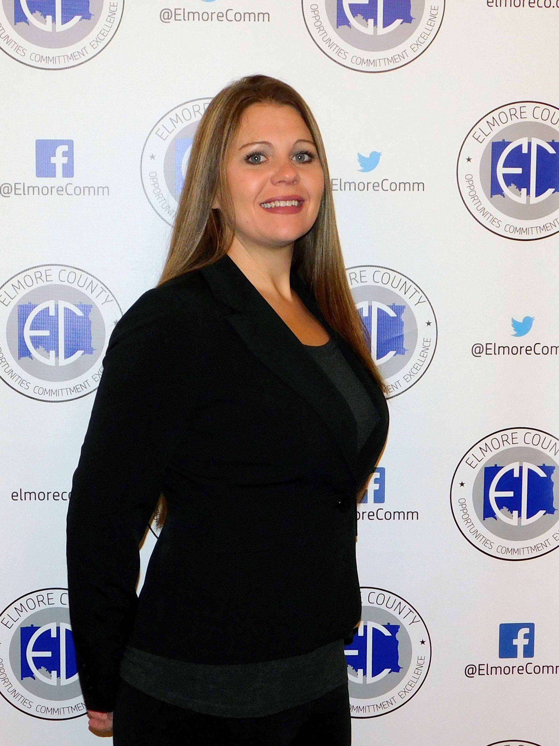 Woman with long brown hair, in black blazer, stands in front of a wall with blue and white logo pattern.