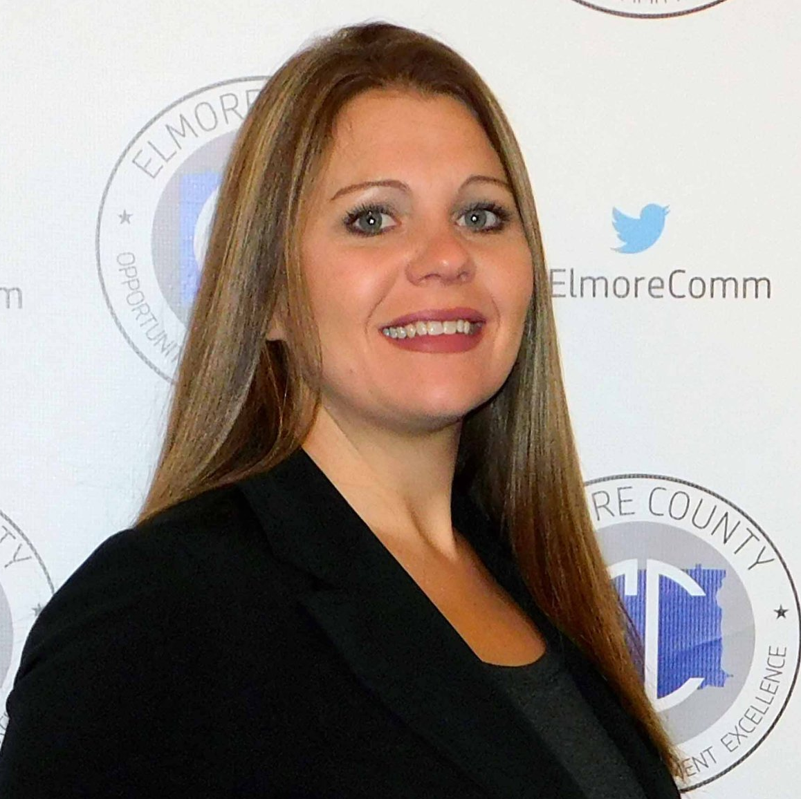 Woman with long brown hair smiles at the camera, wearing a black blazer. The background has logos for Elmore County, Alabama.