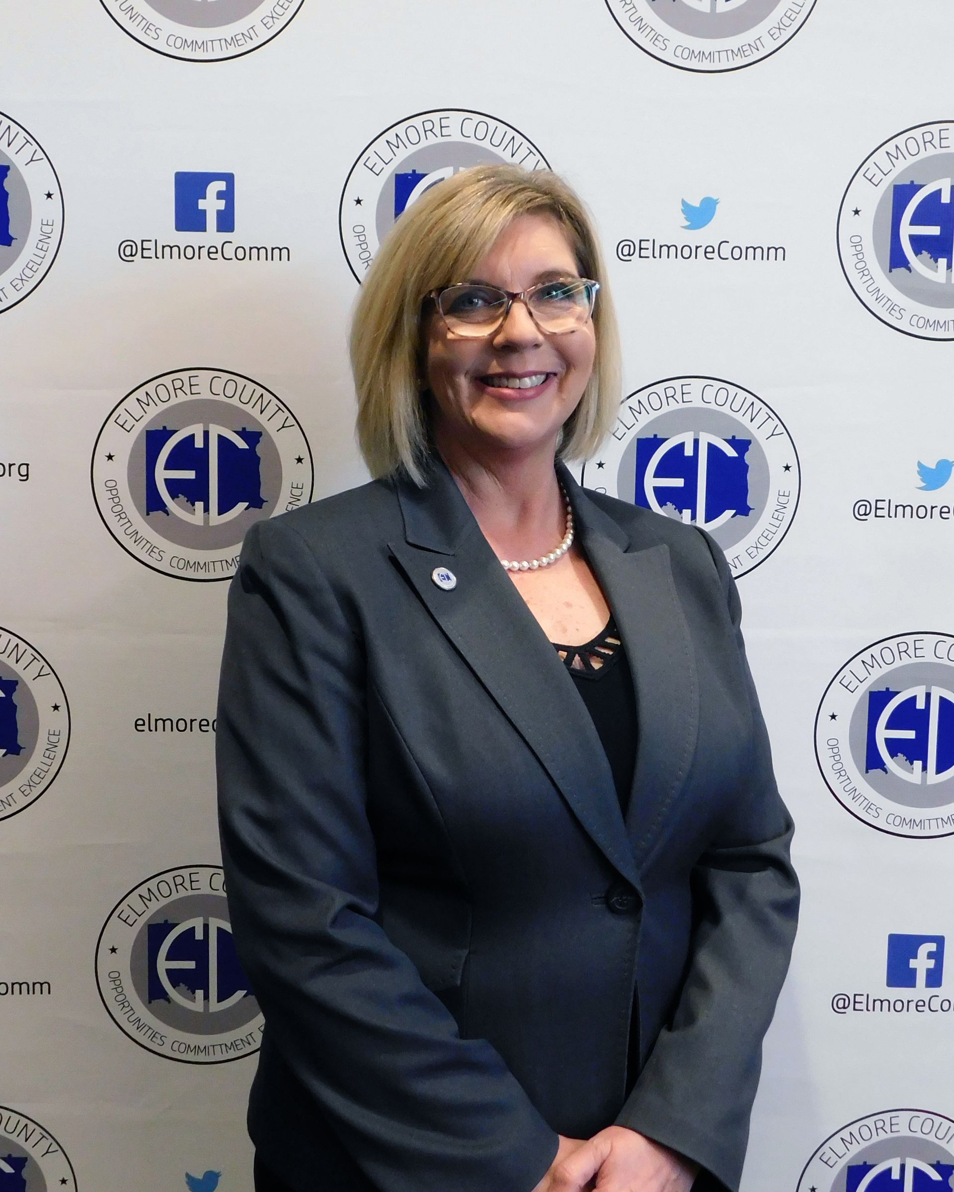 Woman in a gray blazer, smiling, wearing glasses, standing in front of a backdrop with a logo.