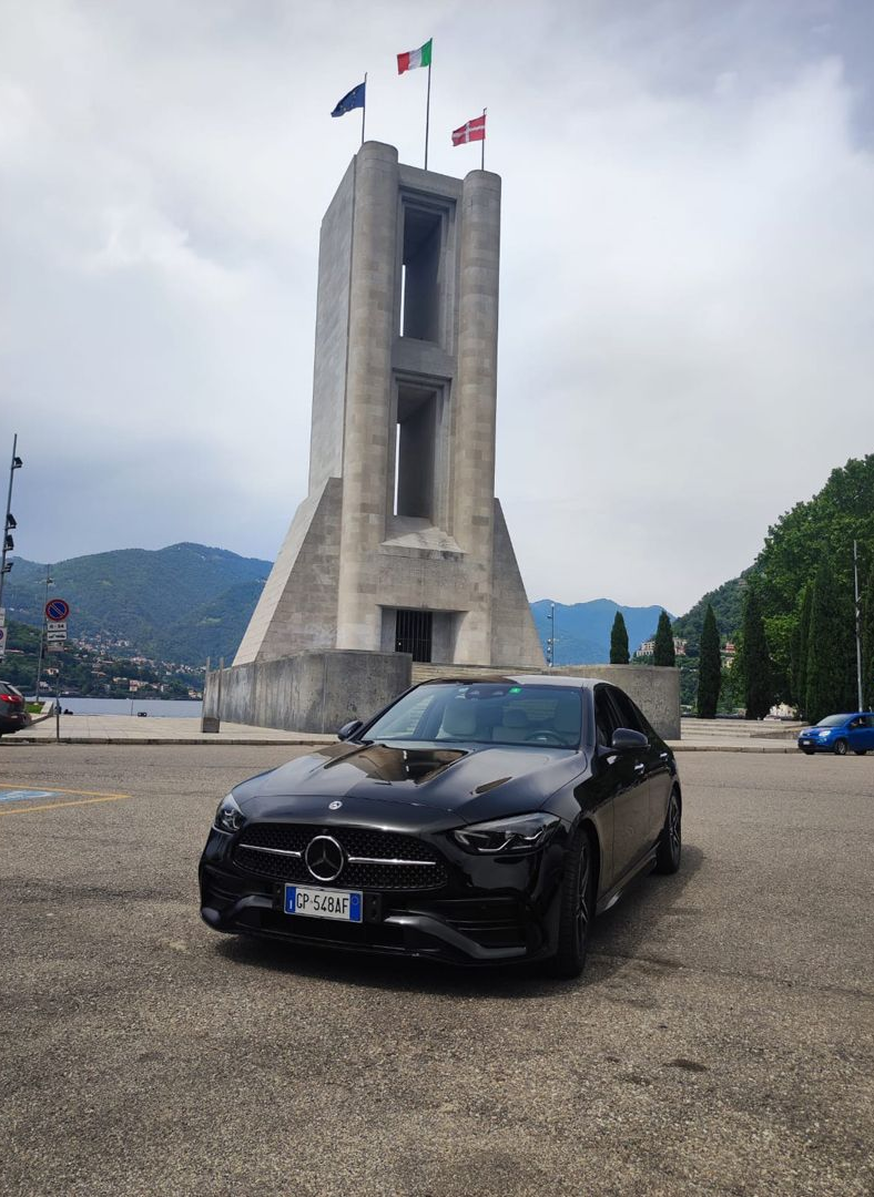 Una berlina Mercedes-Benz nera parcheggiata davanti a un monumento in pietra con bandiere, con vista su un lago e sulle montagne.