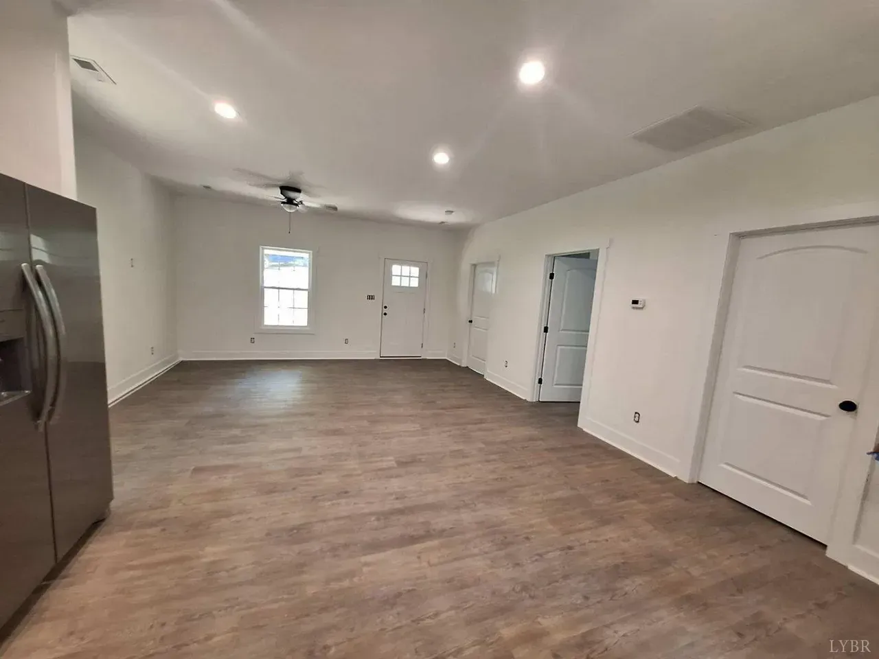 An open-plan room with wood-look flooring, white walls, recessed lighting, two closed doors, and a stainless steel fridge.