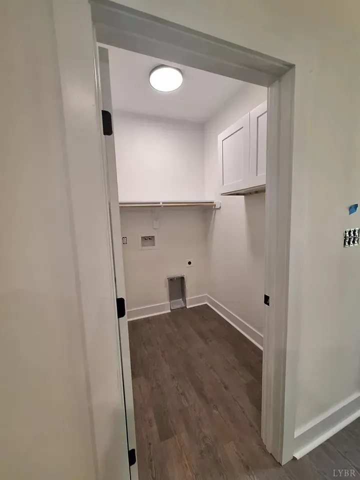 A small, empty laundry room with light walls, wood-look flooring, overhead cabinets, a shelf, and washer/dryer hookups.