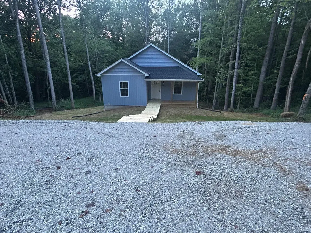 A light blue, single-story house with a grey roof sits in a wooded area, accessed by a path over a large gravel lot.